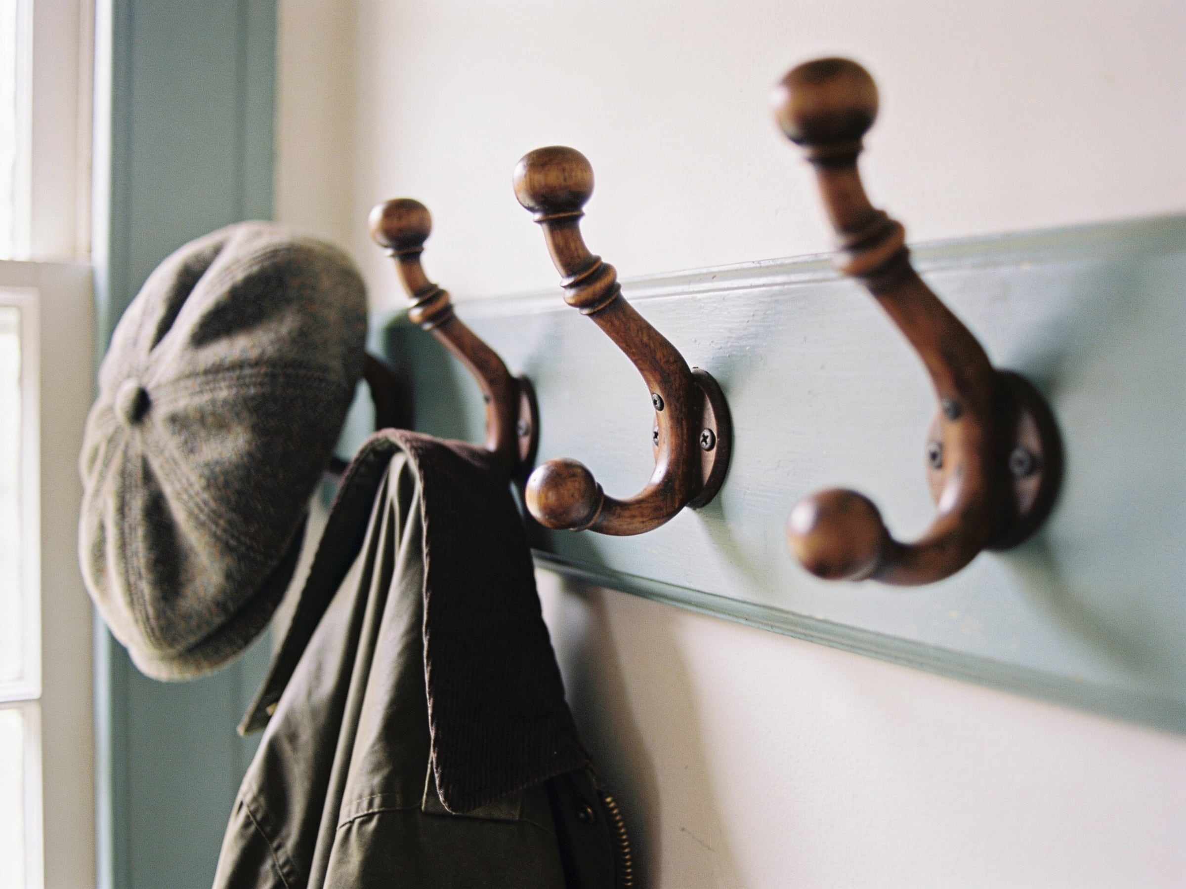 Close-up of three curved wooden coat hooks on a pale painted rail, a tweed cap and a waxed jacket hanging from them
