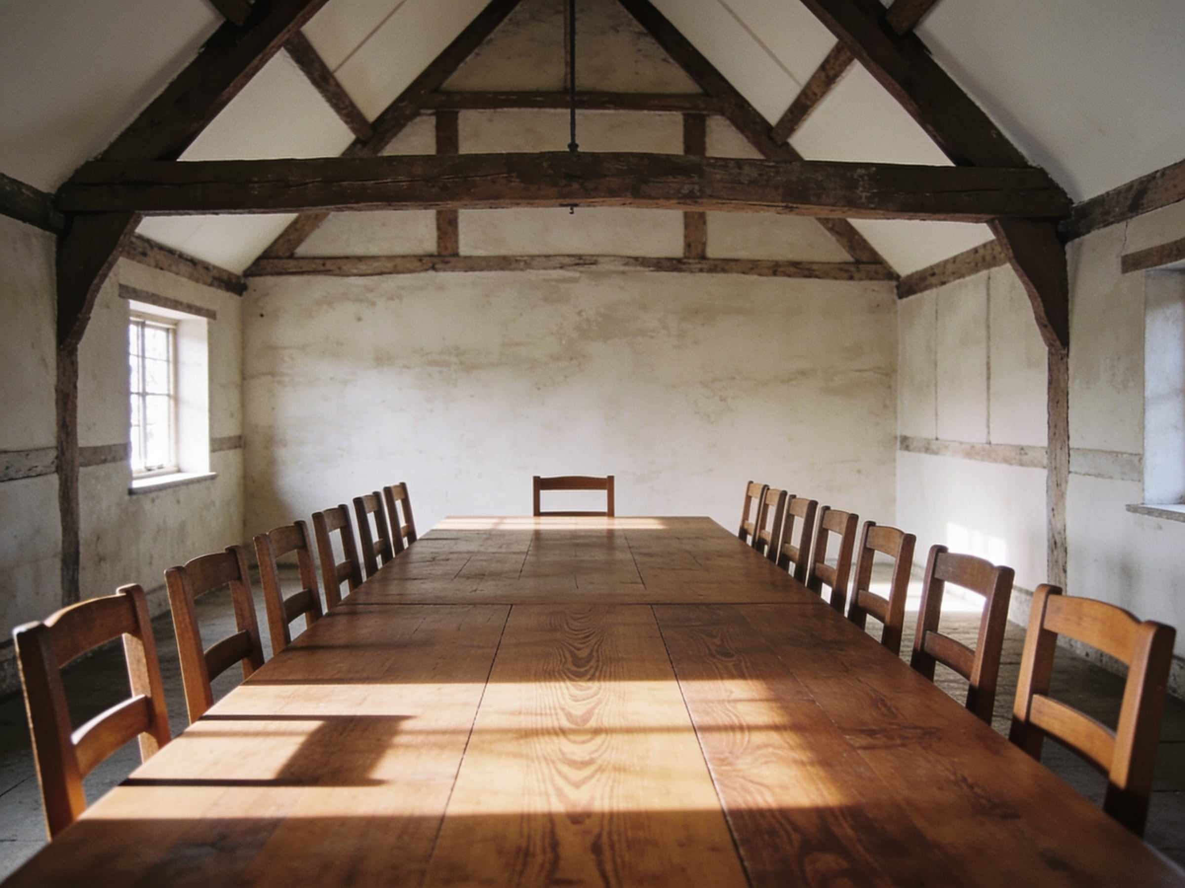 A long wooden dining table with twelve simple wooden chairs in a whitewashed barn with old oak trusses, low winter sun across the table top