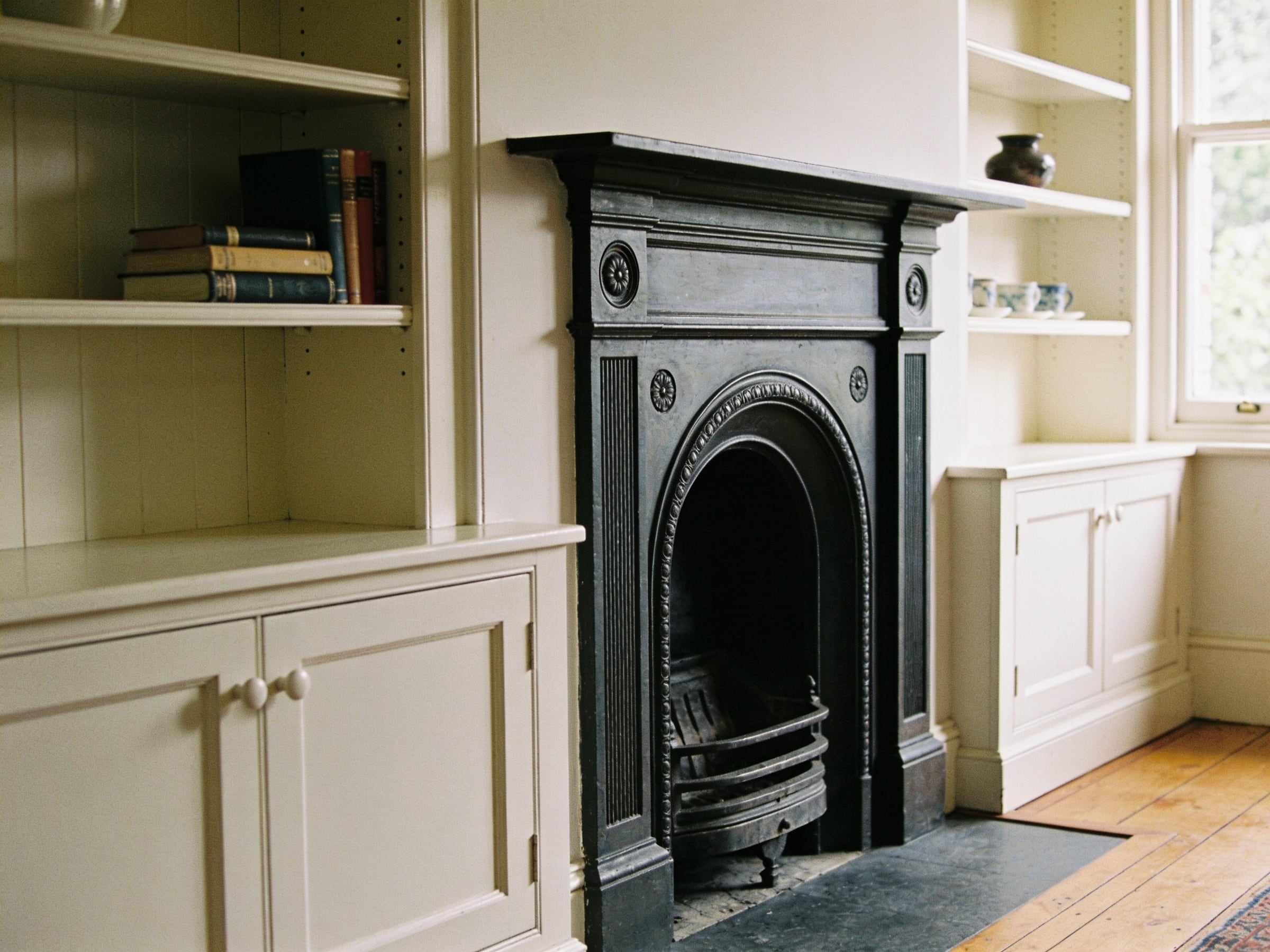 Painted cream alcove cupboards and open shelves either side of a black cast iron fireplace, with a few books and a vase on the shelves